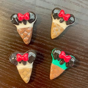 Può includere: Quattro piccoli cabochon in resina a retro piatto a forma di coni gelato sormontati da orecchie di topo nere e un fiocco rosso brillante. Ogni pezzo presenta una base a cono testurizzata in tonalità marrone o beige, con orecchie nere lucide.