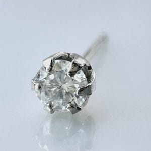 Puede incluir: Primer plano de un pendiente de diamante único. El diamante de talla redonda está engastado en una montura de plata con garras. El poste del pendiente es visible desde la parte posterior. El fondo es blanco y borroso. Una joya clásica.