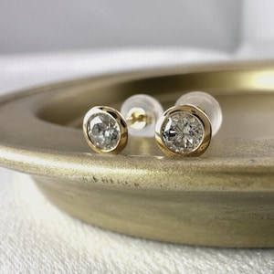 Op de afbeelding: Een paar gouden diamantstud-oorbellen met bezel-zetting. De ronde diamanten zijn gezet in een gepolijste gouden bezel en de oorbellen worden gepresenteerd op een goudkleurige schaal. De oorbellen worden getoond met hun doorzichtige oorbelruggen.