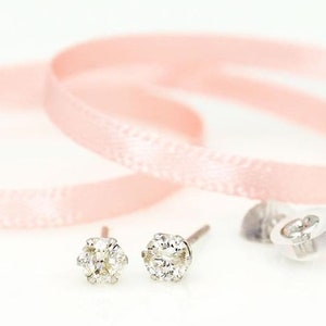 May include: A pair of diamond stud earrings with a six-prong setting, displayed on a white surface. The earrings are next to a pink ribbon, and a clear earring back is visible. The earrings are a classic jewelry item.
