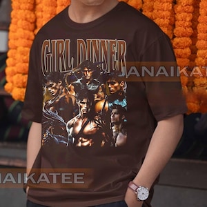 May include: Brown t-shirt with the text "GIRL DINNER" in a vintage style font. The shirt features a collage of muscular male figures. The shirt is a crew neck and has short sleeves.