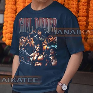 May include: Navy blue t-shirt with the text "GIRL DINNER" in a vintage style. The shirt features a collage of muscular men in various poses. The overall design has a retro aesthetic.
