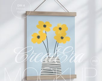 Può includere: Una stampa artistica incorniciata con un'illustrazione floreale. L'opera presenta fiori gialli con steli neri in un vaso a strisce su uno sfondo azzurro. La cornice è in legno chiaro ed è sospesa da una corda nera.