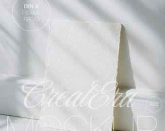 May include: A white rectangular paper with a textured surface, leaning against a white wall. The paper has a deckled edge. The image includes the text "DIN A FRAME RATIO" and "CreateEra MOCKUP".