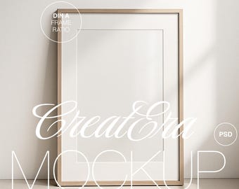 Può includere: Un mockup di una cornice in legno chiaro con sfondo bianco. La cornice è appoggiata su una superficie beige chiaro. Il testo "Create a Mockup" è scritto in lettere bianche in corsivo nella parte inferiore dell'immagine. Il testo "DIN A FRAME RATIO" è scritto in bianco all'interno di un cerchio nell'angolo in alto a sinistra dell'immagine.