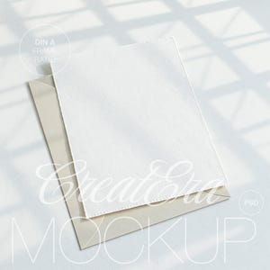 May include: A white textured paper with a deckled edge rests atop a light beige envelope. The image is a mockup with the text "DIN A FRAME RATIO" and "Create Era MOCKUP" in the background.