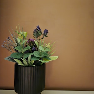 May include: A black ceramic planter with a ribbed design holds a bouquet of artificial flowers. The flowers are a mix of green and purple.