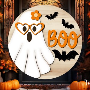 May include: A wooden round sign with a white ghost wearing glasses and a flower on its head. The sign says "BOO" in orange letters and has black bats around the ghost.