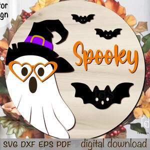 Pode incluir: Decora&ccedil;&atilde;o redonda de Halloween em madeira com um fantasma branco usando um chap&eacute;u de bruxa e &oacute;culos em forma de cora&ccedil;&atilde;o. A palavra "Spooky" &eacute; laranja, com silhuetas de morcegos pretos. Folhas de outono no fundo.