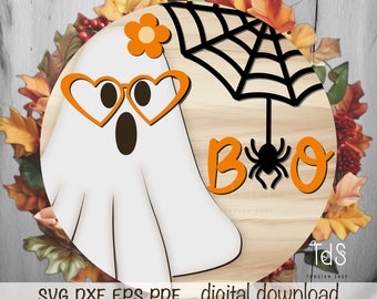 May include: A round wooden sign with a white ghost wearing orange heart-shaped sunglasses and a flower on its head. The ghost is on the left side of the sign. On the right side of the sign is a black spider web with a spider hanging from it. The words "BOO" are written in orange letters below the spider web.