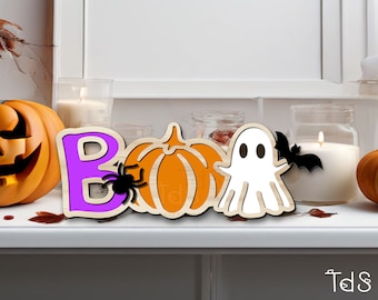 May include: Wooden Halloween decoration spelling "BOO" with a purple letter B, orange pumpkin, white ghost, and black spider and bat. The decoration is on a white surface with lit candles and a carved pumpkin in the background.