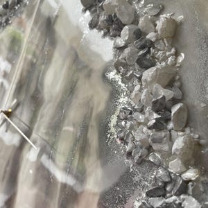 May include: A close-up of a gray and white resin clock face with a silver clock hand. The clock face is decorated with small, white and gray stones.