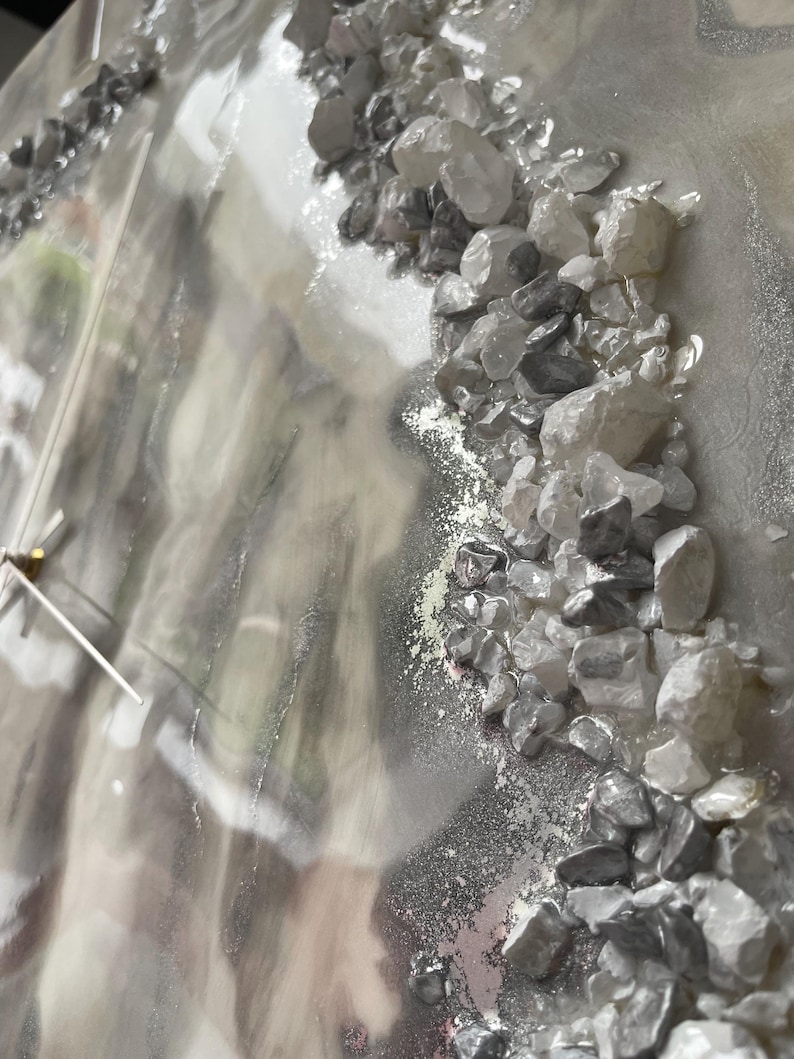 May include: A close-up of a gray and white resin clock face with a silver clock hand. The clock face is decorated with small, white and gray stones.