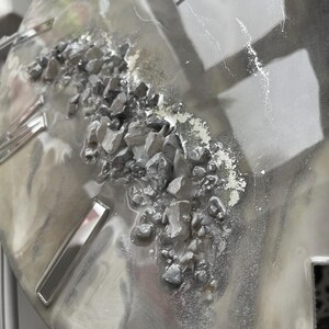 May include: A close-up of a gray and silver resin art piece with a textured surface. The piece features a cluster of small, gray rocks embedded in the resin.