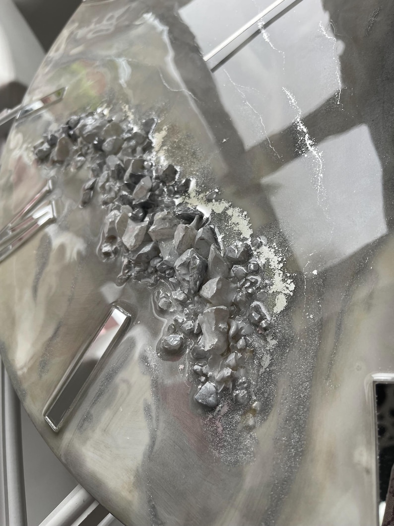May include: A close-up of a gray and silver resin art piece with a textured surface. The piece features a cluster of small, gray rocks embedded in the resin.