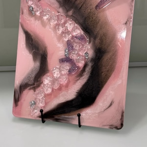 May include: A pink and black resin tray with a swirl design and clear and purple glitter. The tray has a small, clear, pointed object on top.