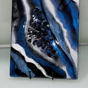 May include: A square resin art piece with a blue, white, and black abstract design. The design features a cluster of black resin pieces in the center of the piece.