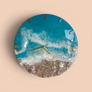 May include: A round wall clock featuring a glossy resin finish that mimics an ocean scene with swirling turquoise and white waves crashing onto a sandy beach. The clock face includes gold-colored Roman numerals and thin gold-colored hands.