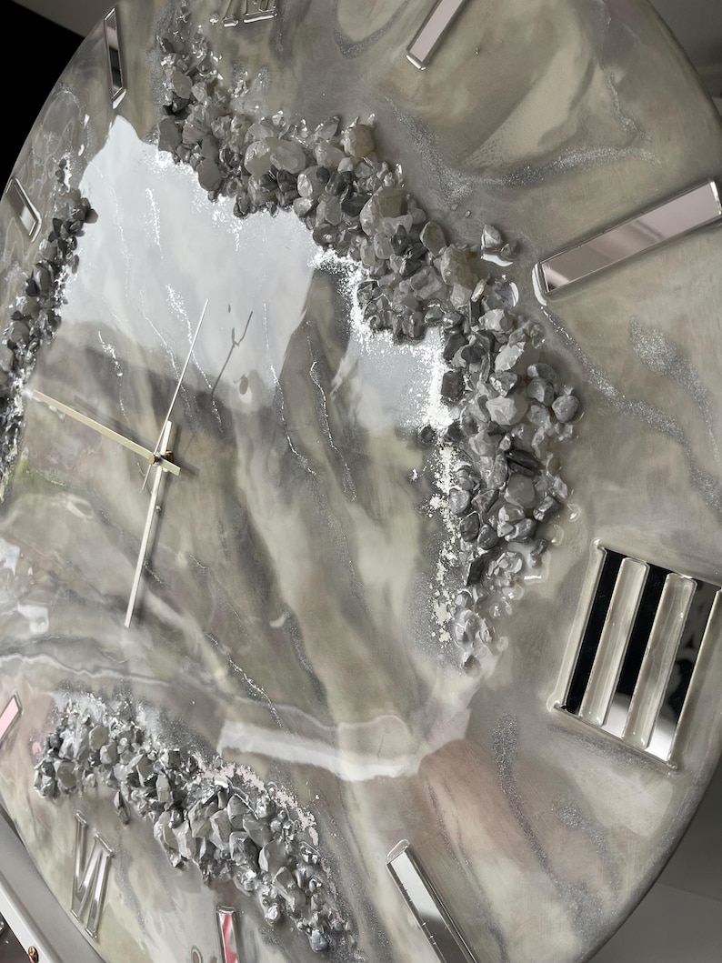 May include: A gray and white resin clock face with silver accents and embedded gray stones. The clock face has Roman numerals and a silver hand.