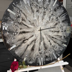 May include: A large, round wall clock with a black and silver resin design. The clock face features Roman numerals and mirrored accents. The clock is displayed on a white easel, with a red rose in the foreground.
