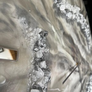 May include: A close-up of a gray and white resin clock face with a silver hand and a silver and white rock design.