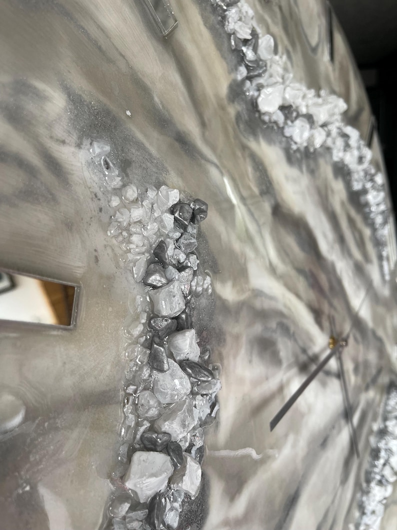May include: A close-up of a gray and white resin clock face with a silver hand and a silver and white rock design.