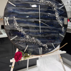 May include: A black and white resin clock with silver Roman numerals and a white clock hand. The clock is on a white easel and has a blue and white marbled design with silver glitter.