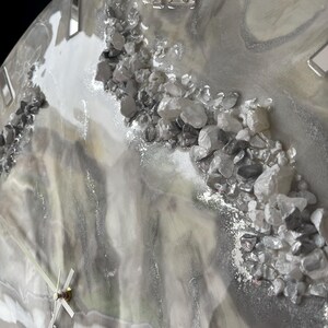 May include: A gray and white resin clock face with Roman numeral hour markers and silver metallic accents. The clock face is decorated with small, clear crystals.