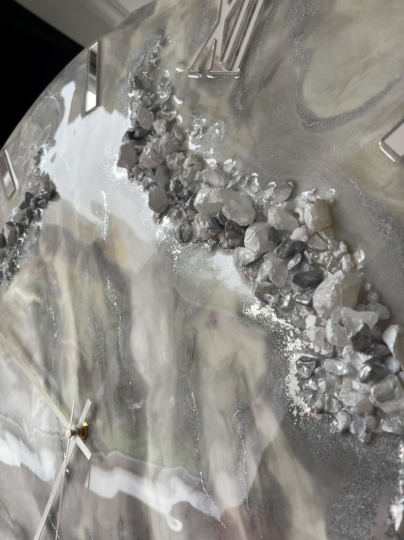 May include: A gray and white resin clock face with Roman numeral hour markers and silver metallic accents. The clock face is decorated with small, clear crystals.