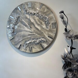May include: A large, round wall clock with a grey, white and hint of beige marble effect resin face. Silver Roman numerals and metallic accents are incorporated into the design.  The clock features embedded, small, shiny, off-white stones or crystals adding texture and visual interest.  The overall style is modern and elegant.