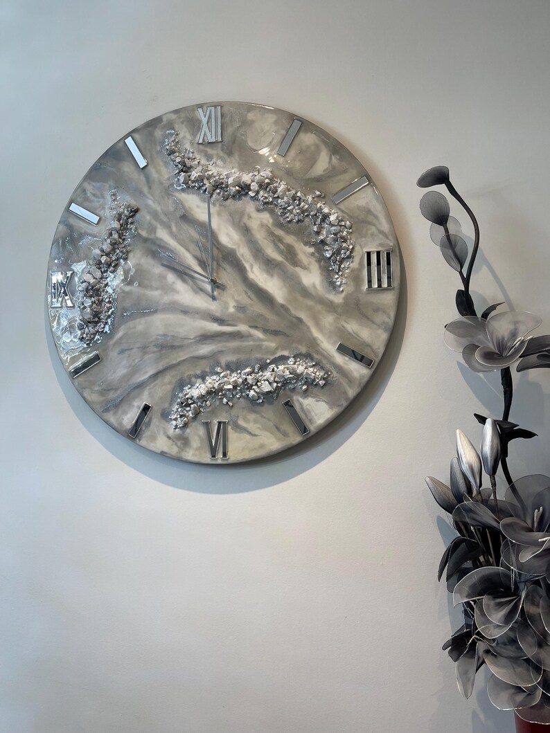 May include: A large, round wall clock with a grey, white and hint of beige marble effect resin face. Silver Roman numerals and metallic accents are incorporated into the design.  The clock features embedded, small, shiny, off-white stones or crystals adding texture and visual interest.  The overall style is modern and elegant.