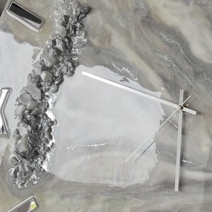 May include: A gray and white resin clock with silver accents and a silver metal frame. The clock face has a marble-like design with silver glitter and small silver rocks embedded in the resin. The clock hands are silver metal.