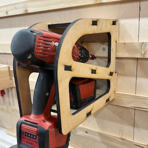 May include: A wooden wall mount for a cordless drill and battery. The mount is made of light brown wood and has a cutout for the drill and battery. The drill is red and black and has a battery attached to it. The battery is red and black and has the text "14.4 Volt Li-Ion 3.3 Ah" on it.