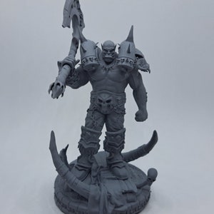 May include: A gray 3D printed miniature of an Orc warrior holding a large axe. The Orc is wearing armor and a belt with a skull buckle. The miniature is standing on a circular base with a skull and bones.