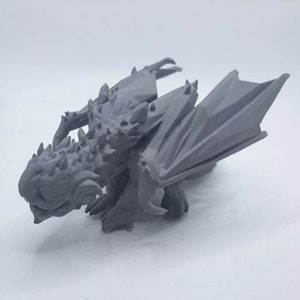 Può includere: Una piccola statuina di drago stampata in 3D, di colore grigio, con le ali spiegate. Il drago ha una schiena spinosa e un viso dettagliato.