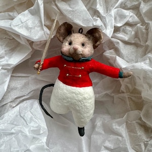 May include: A felt mouse ornament wearing a red jacket with gold buttons, white pants, and black shoes. The mouse holds a silver sword and has large brown ears. The ornament is set against a white, textured background.