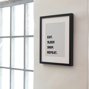 May include: A framed print with a black border displays the words "EAT. SLEEP. SHOP. REPEAT." in a bold, sans-serif font. The print is mounted on a white background and hangs on a white wall, with a window and greenery visible in the background.