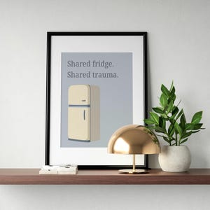 May include: A framed art print featuring a retro refrigerator illustration in beige and gray, with the text "Shared fridge. Shared trauma." The print is displayed on a brown shelf with a gold lamp and a potted plant.