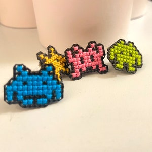 Puede incluir: Cuatro parches bordados de pixel art con invasores espaciales en azul, rosa, amarillo y verde.