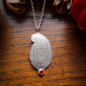 Peut inclure: Un collier pendentif argenté orné d'un charm en forme de paisley, avec des détails en relief. Une petite pierre précieuse rouge et ronde est incrustée à la base du charm. La chaîne en argent s'accorde avec le pendentif. Présenté sur une surface en bois.