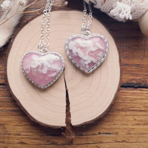 Peut inclure: Deux pendentifs en forme de cœur avec des chaînes argentées. Les pendentifs présentent un fond rose avec des motifs de nuages blancs et sont encadrés d'une bordure argentée perlée. Ils reposent sur une tranche de bois, avec des fleurs séchées en arrière-plan.