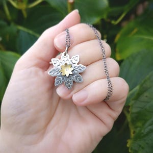 Op de afbeelding: Een zilveren en gouden ketting met een bloemhanger. De hanger is gemaakt van twee lagen metaal, waarbij de bovenste laag van goud is en de onderste laag van zilver. De bloem heeft een kleine diamant in het midden.