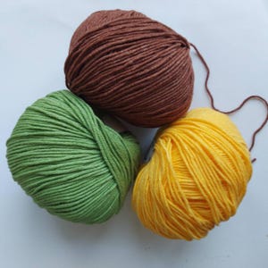 May include: Three round skeins of yarn arranged in a cluster. One skein is a deep brown, one is a vibrant grass green, and one is a bright yellow. Each skein features a smooth, tightly wound texture with a single loose strand extending from the brown yarn.