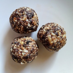 May include: Three round energy bites on a white plate. The bites are a mix of brown, tan, and white, with visible nuts and dried fruit. The texture appears to be dense and slightly coarse, suggesting a homemade or natural food product.