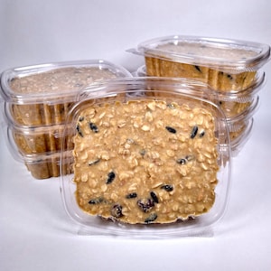 May include: Several clear plastic containers filled with birdseed cakes. The cakes are rectangular and have a light brown color with visible seeds and grains. The containers are stacked, suggesting multiple units of the product.