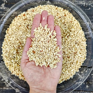 May include: A clear glass bowl filled with small, light brown, irregularly shaped pieces. A hand is cupped, holding a portion of the pieces. The bowl sits on a dark, weathered wooden surface.