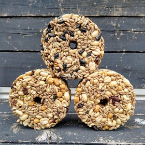 May include: Three round bird feeders made of pressed seeds, nuts, and berries. The feeders are stacked on top of each other on a dark wooden surface.