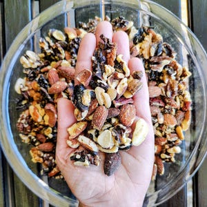 May include: A clear glass bowl filled with a mix of nuts, including almonds, walnuts, and pecans. A handful of the nut mix is held in a hand, showcasing the variety of textures and colors, from light tan to dark brown.