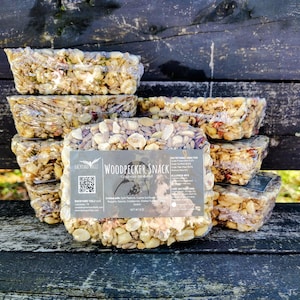 May include: A stack of individually wrapped birdseed bars. The bars are a mix of nuts and seeds. The label reads "Woodpecker Snack" and includes a graphic of a bird. The bars are on a weathered wooden surface.