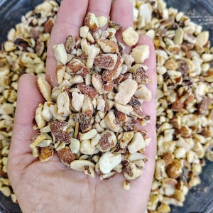May include: A handful of chopped almonds, showcasing a mix of light beige and brown pieces. The nuts are held in an open hand, with more almonds visible in a clear bowl in the background. The image highlights the texture and color variations of the chopped nuts.